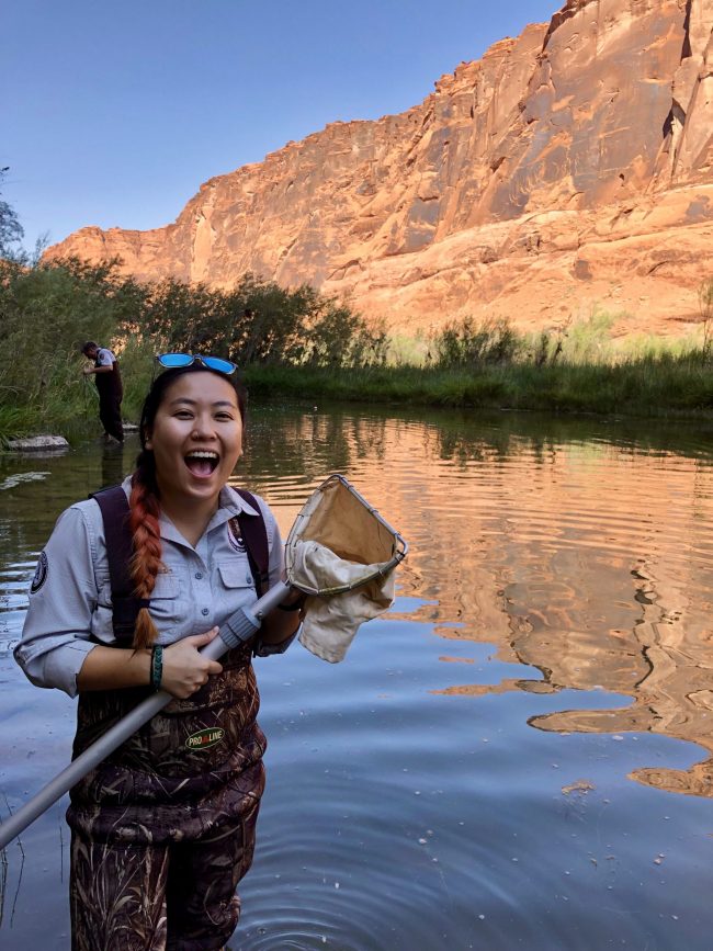 A scienctist stands in knee deep water in a red rock canyon holding a net and a big smile on her face with a